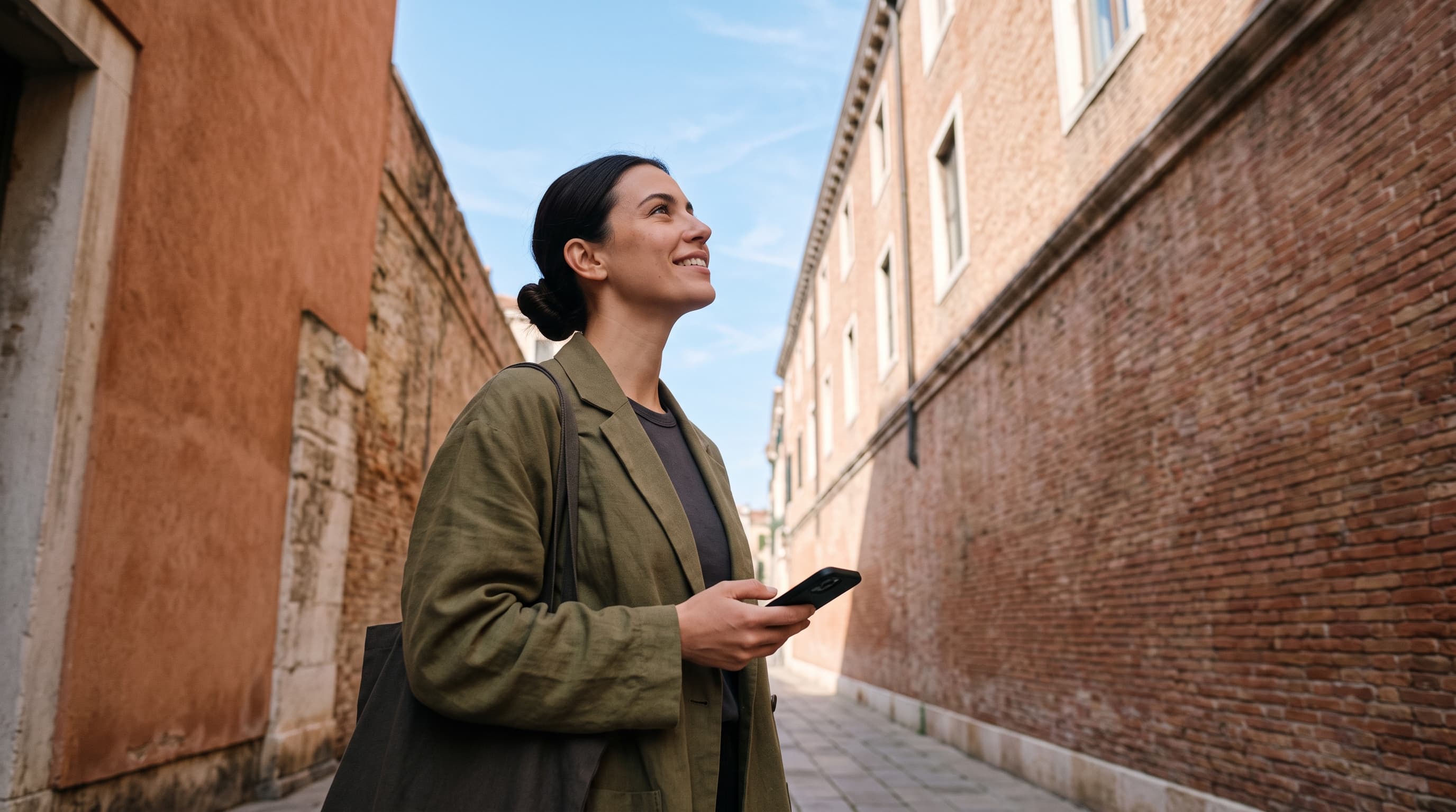A woman in an olive jacket looks calmly up at the sky between a terracotta wall and a brick building